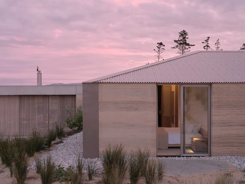 Rammed earth walls and corrugated metal roof among gravel beds and native grasses at dusk