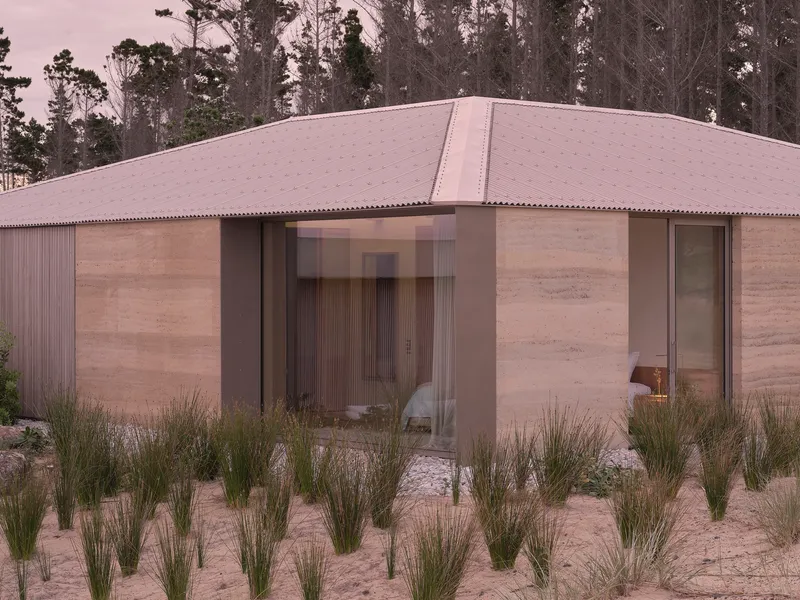 Rammed earth and corrugated metal facade with glazed openings facing sparse grass planting in sandy soil