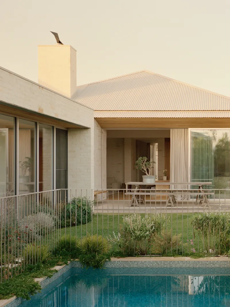 Pool terrace with native grasses and cream-painted masonry walls under a corrugated metal roof at dusk