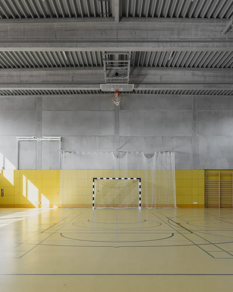 Gymnasium interior with basketball hoop, goal net, exposed concrete ceiling, and yellow wall panels with sunlight