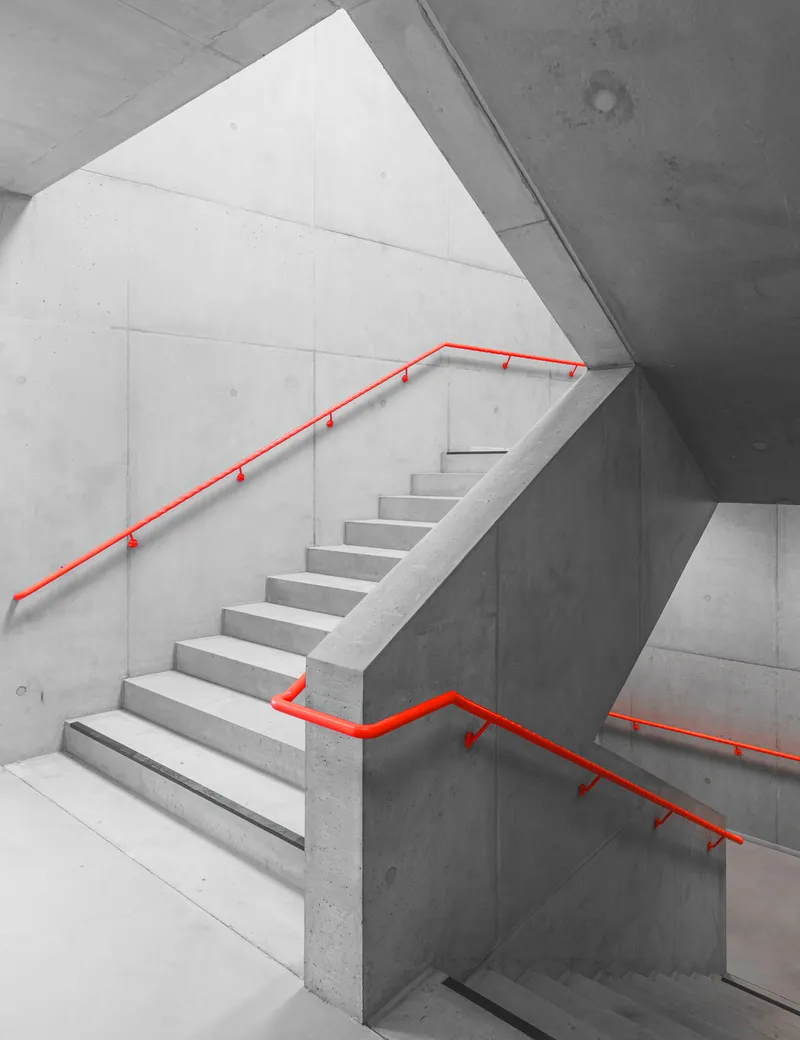 Concrete stairwell with red metal handrails wrapping around angular flights under triangular skylight