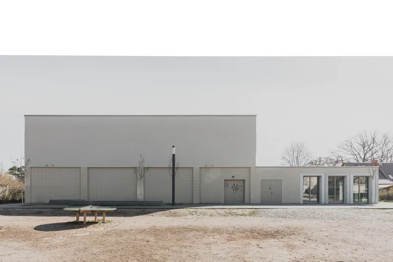 Street view of the grey corrugated metal facade with a single bench on gravel under bare winter trees