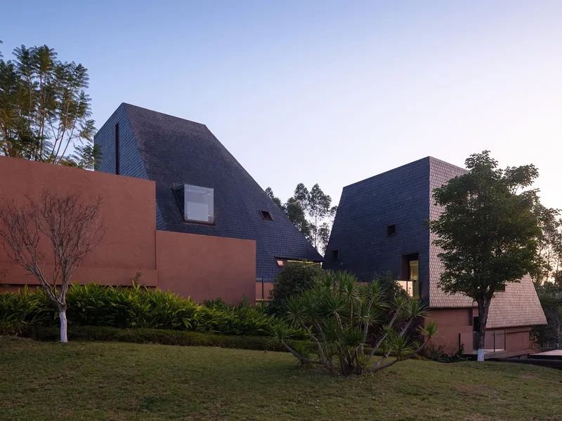 Clustered volumes clad in dark shingles rising above planted lawn and terracotta walls at dusk