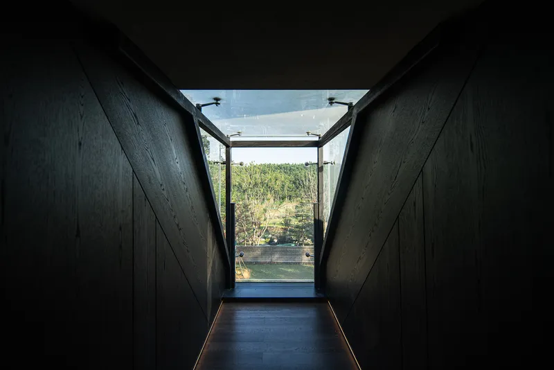 Dark timber-lined corridor with angled walls opening to glass door and landscape beyond