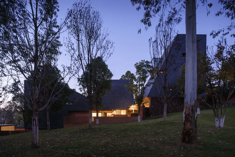 Night view of the low-slung residence with illuminated windows behind bare winter trees on sloping lawn
