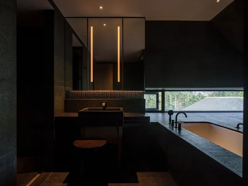 Dark bathroom vanity with vertical strip lighting above twin sinks and horizontal window overlooking treetops