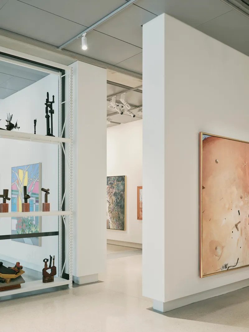 Gallery interior with white partition walls displaying artworks on metal shelving and hung on plywood panels