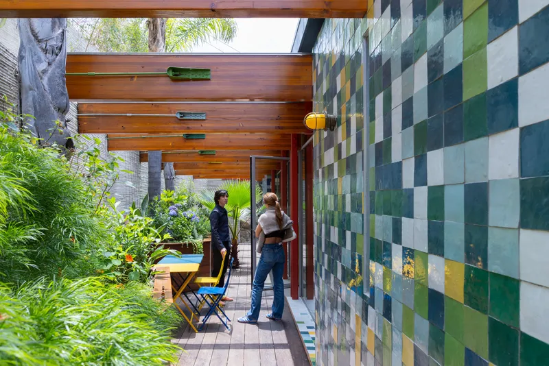 Outdoor corridor with multicolored ceramic tile wall and timber ceiling where two people converse near plantings