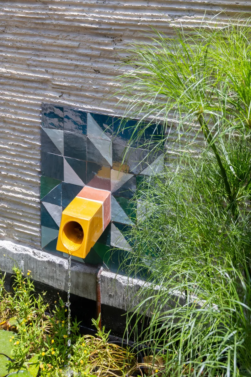 Yellow geometric birdhouse mounted on a checkered tile wall surrounded by tall grasses
