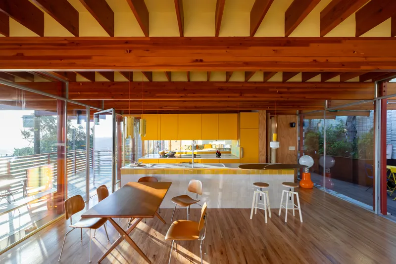 Open-plan kitchen and dining area with exposed timber ceiling joists and glass walls at dusk