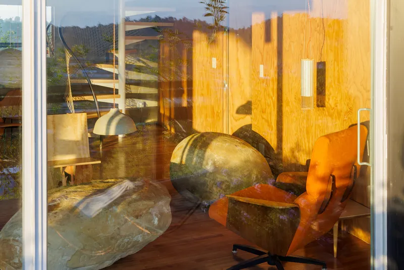 Glass reflection showing interior space with large sculptural stone forms and timber wall panels at sunset
