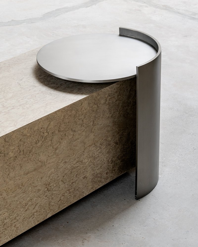 Detail of curved metal edge wrapping a circular table top above a concrete plinth