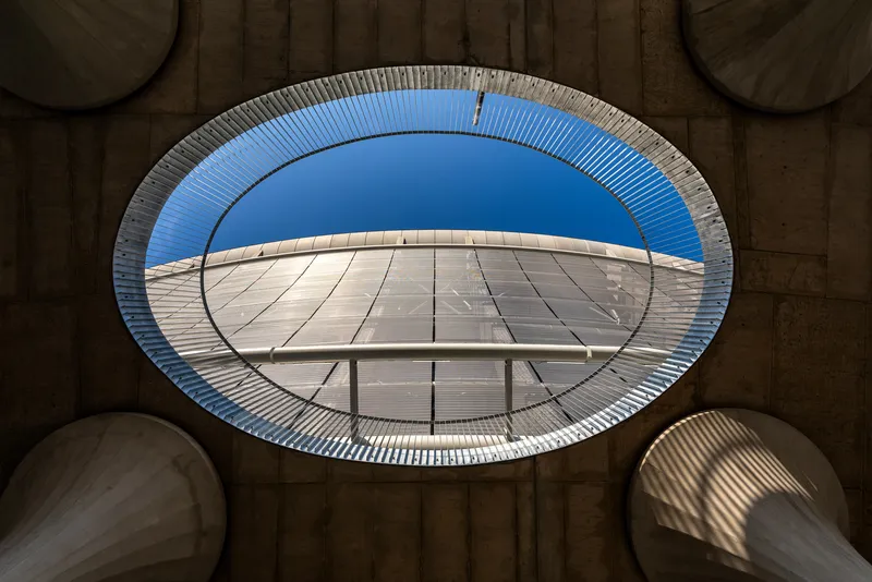 Upward view through an elliptical concrete oculus framing a curved glass canopy and blue sky
