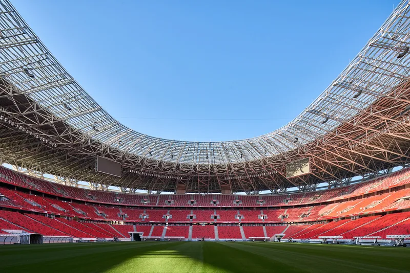 Curved steel truss roof structure spanning over red seating sections and the green striped playing field