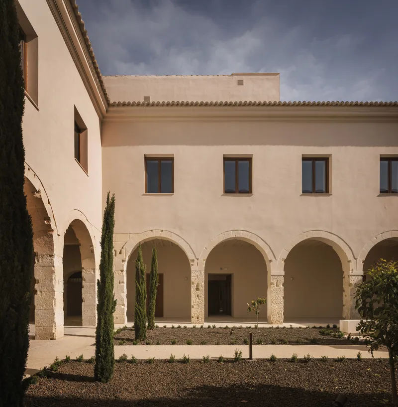 Courtyard facade with stone arcades, plastered upper level, cypress trees, and gravel planted beds below