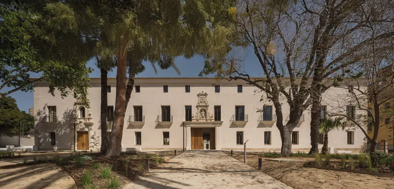 Street facade with ornamental stone portal framed by mature trees casting shadows on gravel forecourt