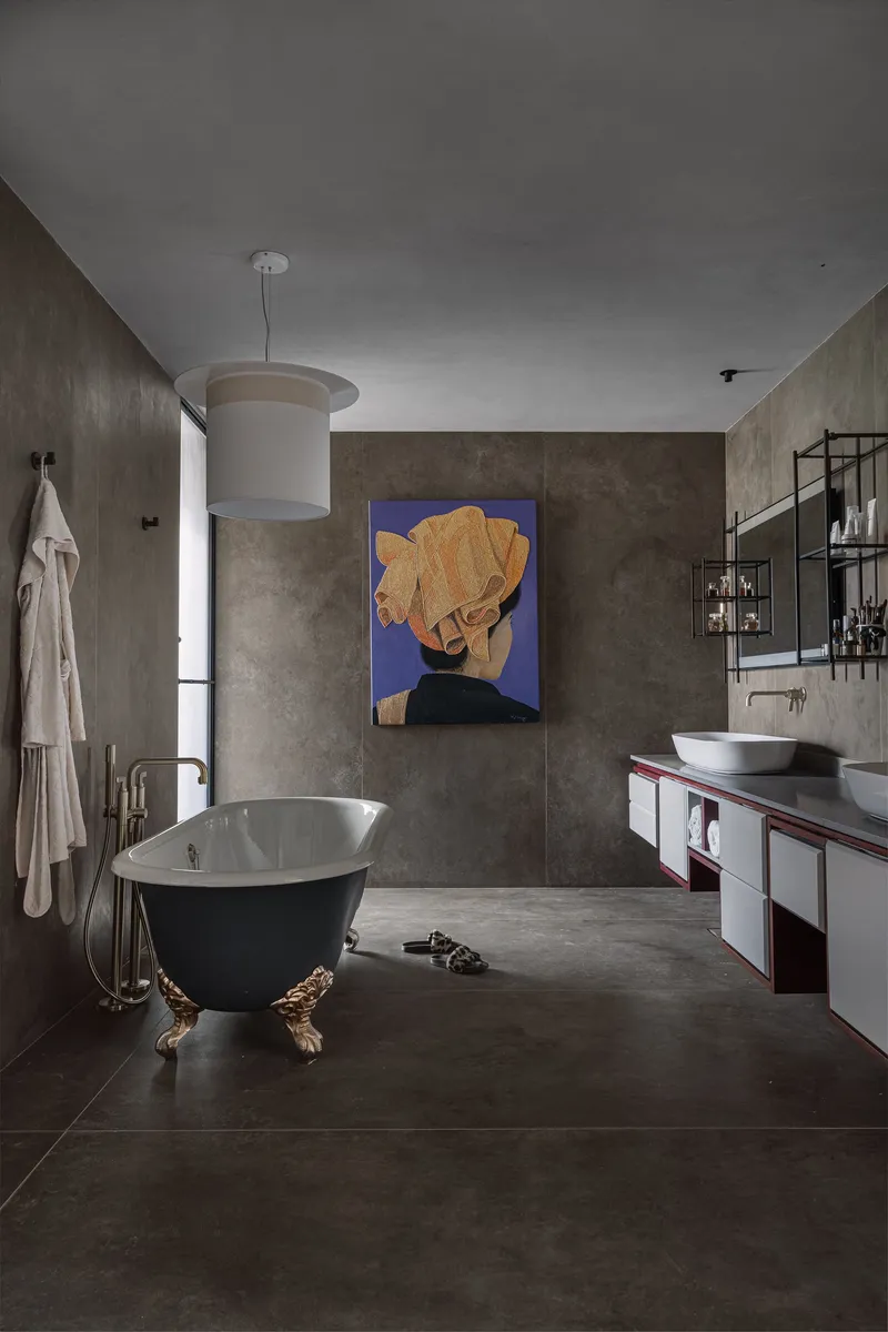 Bathroom with freestanding black tub on brass feet, concrete walls, and abstract portrait painting above