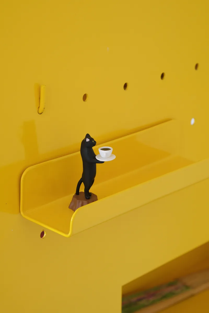 Yellow wall-mounted shelf with a miniature figure holding a cup beneath scattered wall hooks
