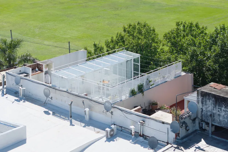 Aerial view of a glass pavilion addition atop a white-painted rooftop adjacent to surrounding green fields