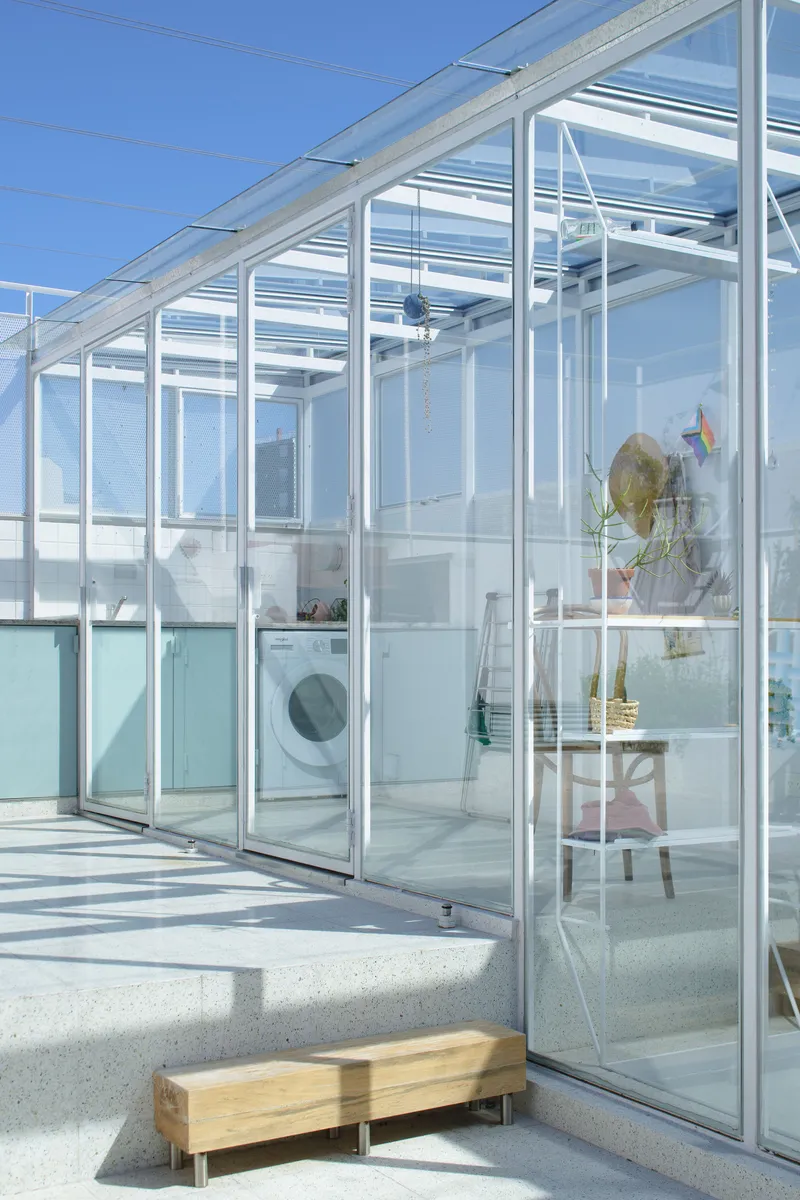 Transparent greenhouse structure with sliding panels and a washing machine visible inside beside a wooden bench