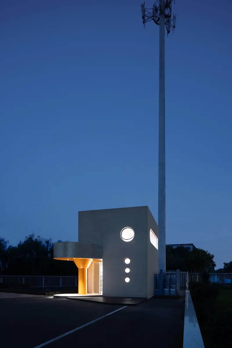 White cubic pavilion with circular portal windows and orange column lit at dusk beside a communications tower