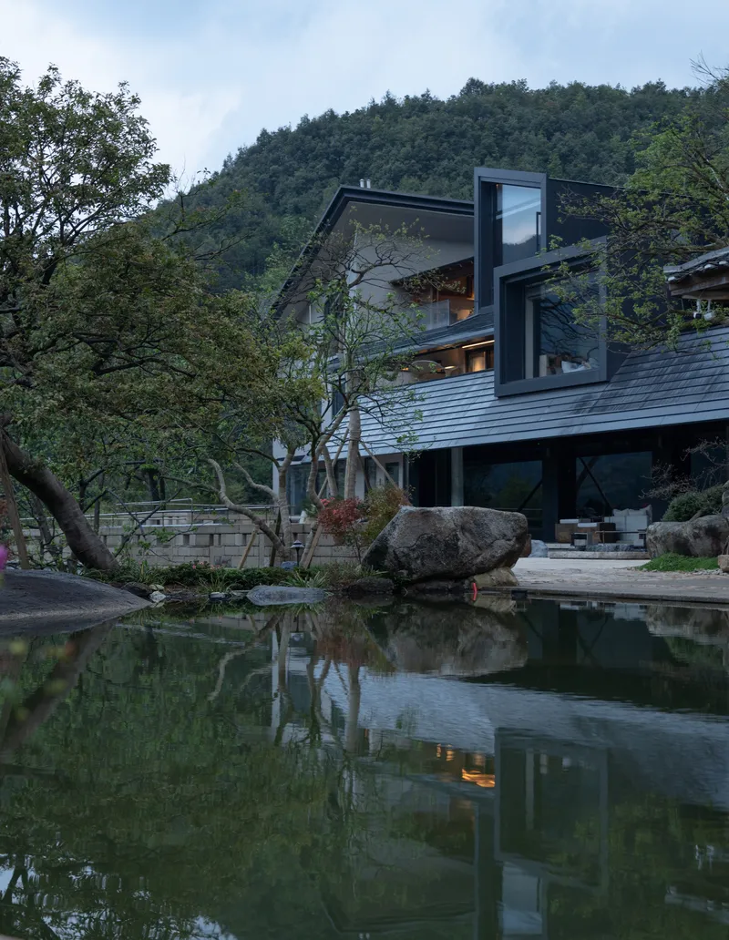 Angled black-framed glazing above pond with boulders and tree reflections at twilight