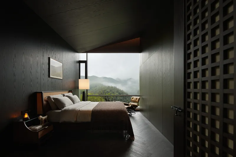 Bedroom with textured dark walls and clerestory window framing mist-shrouded peaks beyond a lounge chair
