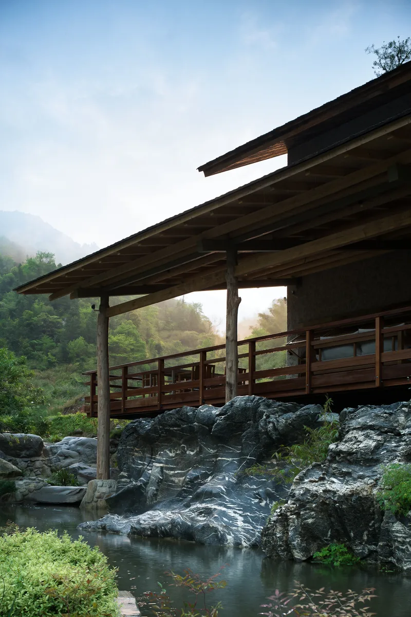 Timber pavilion with exposed rafters spanning a rocky stream in morning mist