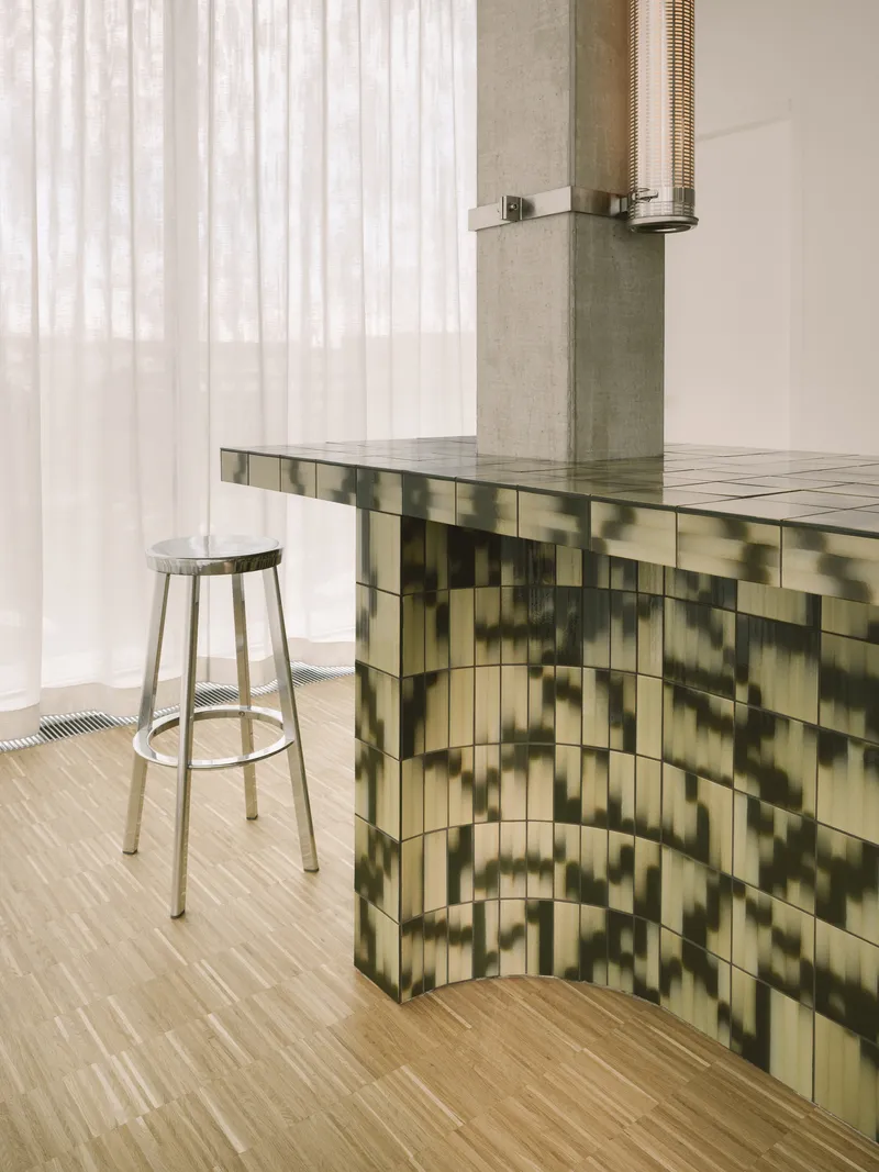 Reception counter clad in green gradient glazed tile with metal stool and sheer curtain backdrop