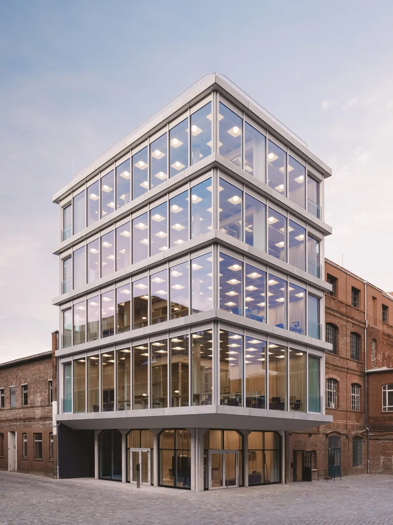 Corner view of the fully glazed facade with white frame and illuminated ceiling grid at dusk