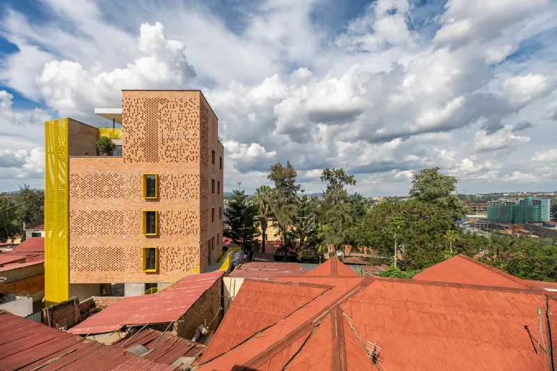 Perforated brick facade with yellow window frames set among trees and low-rise red-roofed buildings