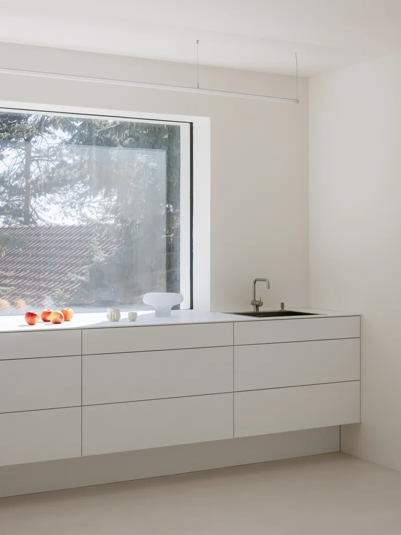 White handleless kitchen cabinetry beneath a horizontal window overlooking pine trees in soft light