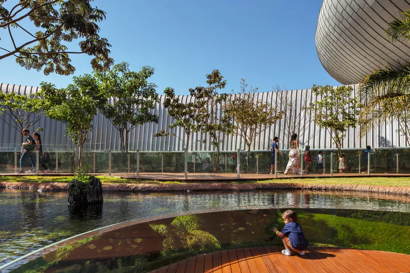 Child sitting on timber deck beside reflecting pool with glass-backed tree promenade beyond