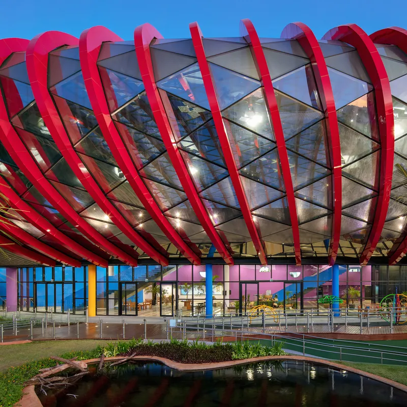 Exterior facade with red arched ribs wrapping glazed envelope above a reflecting pond at twilight