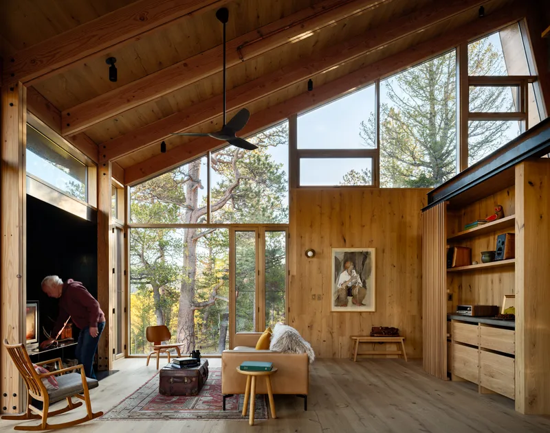 Wood-lined living room with exposed rafters, floor-to-ceiling glazing, and a fireplace with two figures tending it