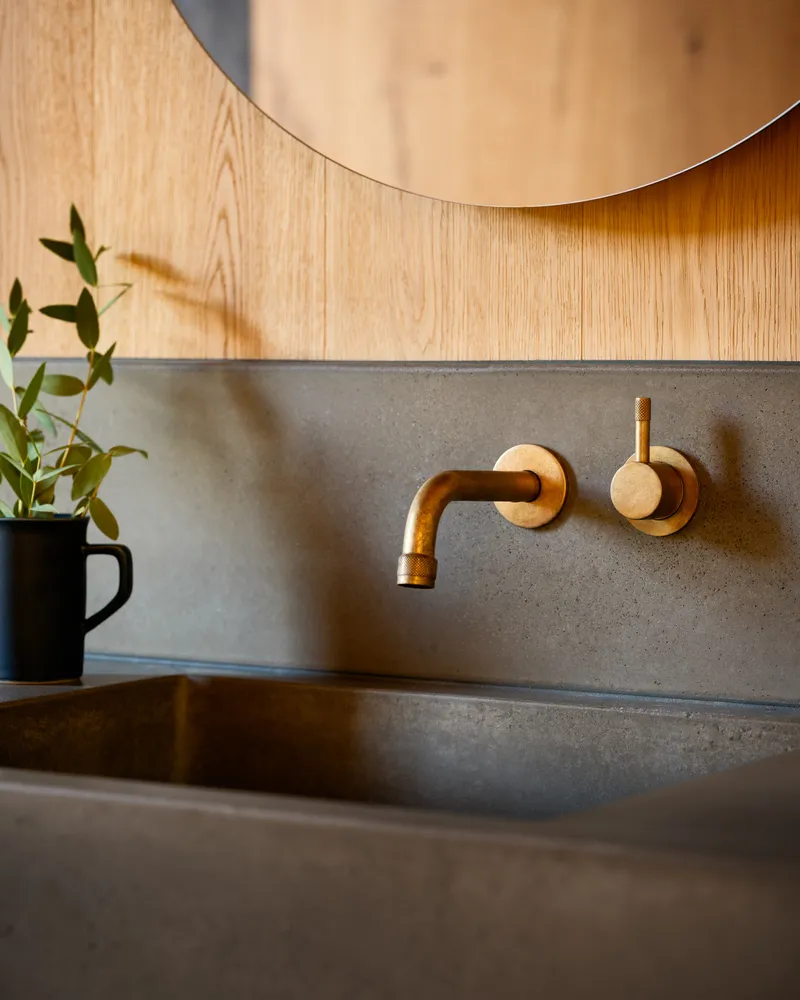 Close-up of brass wall-mounted faucet against concrete backsplash with black mug and greenery