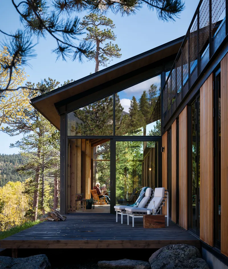 Cantilevered timber and glass volume with deck opening into living room at dusk among pine trees