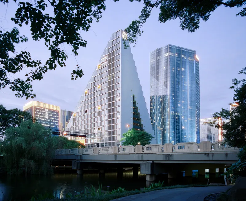 Stepped tower with pixelated facade framed by tree branches and viewed across the river at dusk
