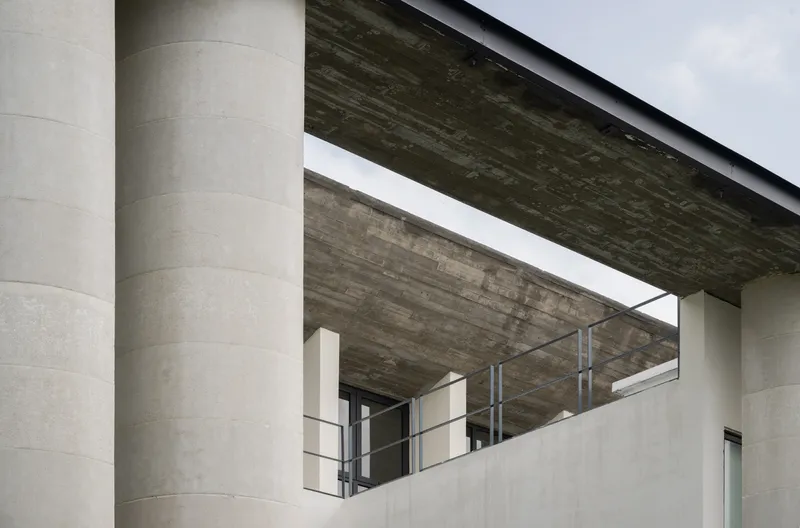 View beneath the cantilevered concrete roof showing cylindrical columns and metal railings under an overcast sky