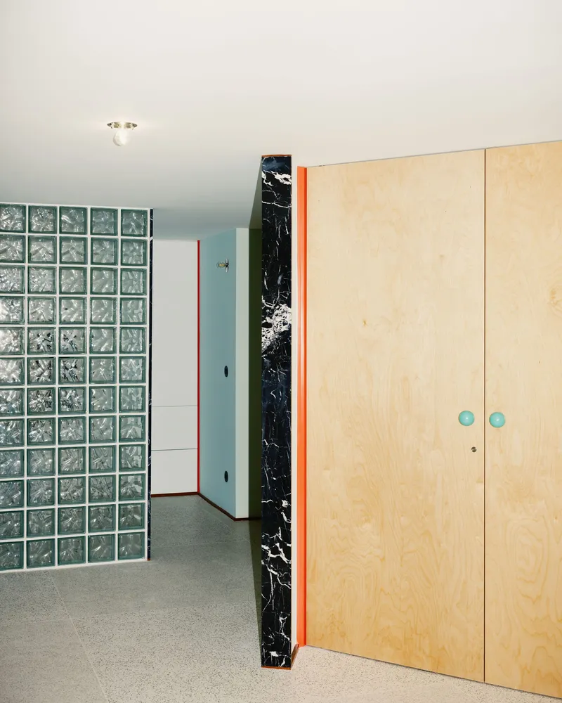 Light plywood wardrobe with blue knobs beside a glass block wall and black marble column