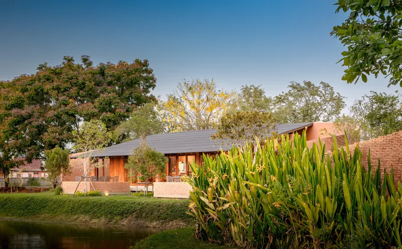 Low-slung pavilion with corrugated metal roof beside a pond edged with ornamental grasses at twilight