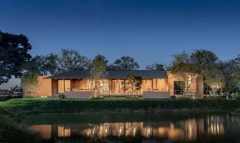 Illuminated single-story pavilion with glass walls reflected in a pond at dusk