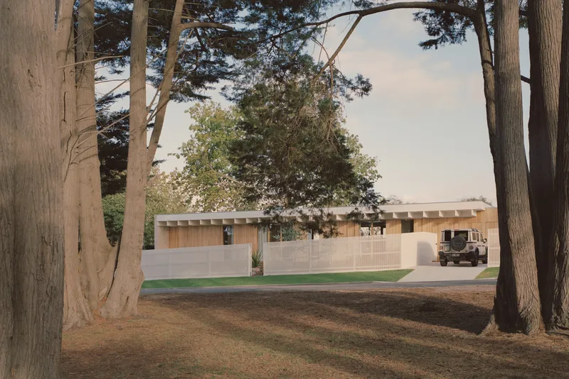 Long cream brick facade with exposed timber rafters viewed through a grove of mature trees at dusk