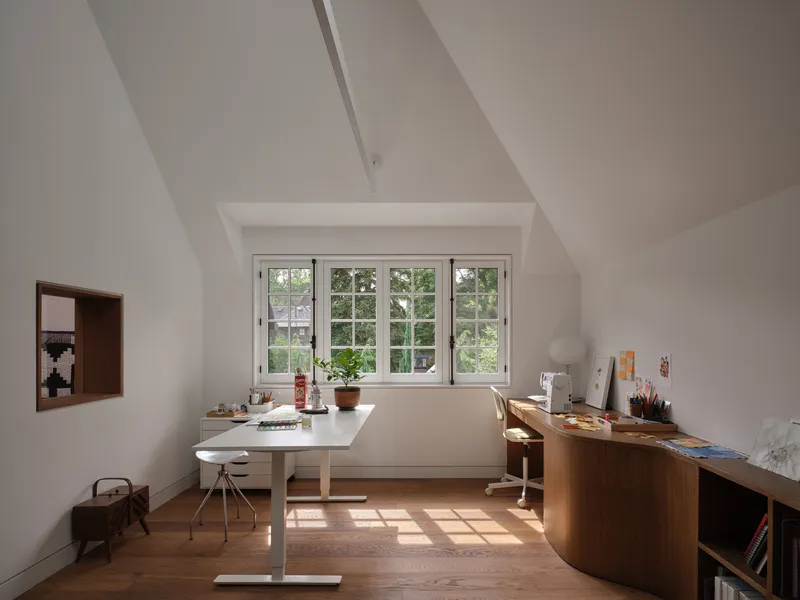 Attic workspace with sloped ceiling, white drafting table, and window overlooking green foliage
