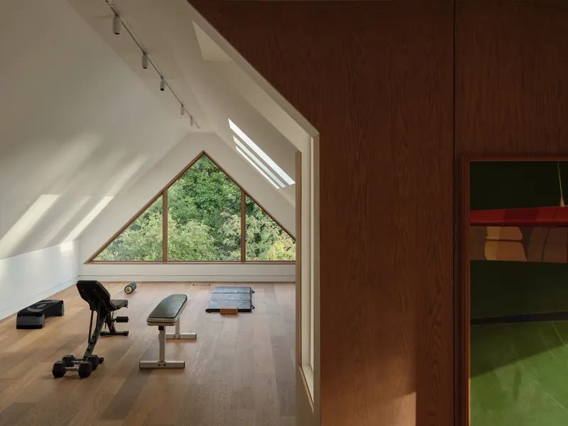 Attic exercise room with triangular window framing green foliage and skylights casting diagonal shadows