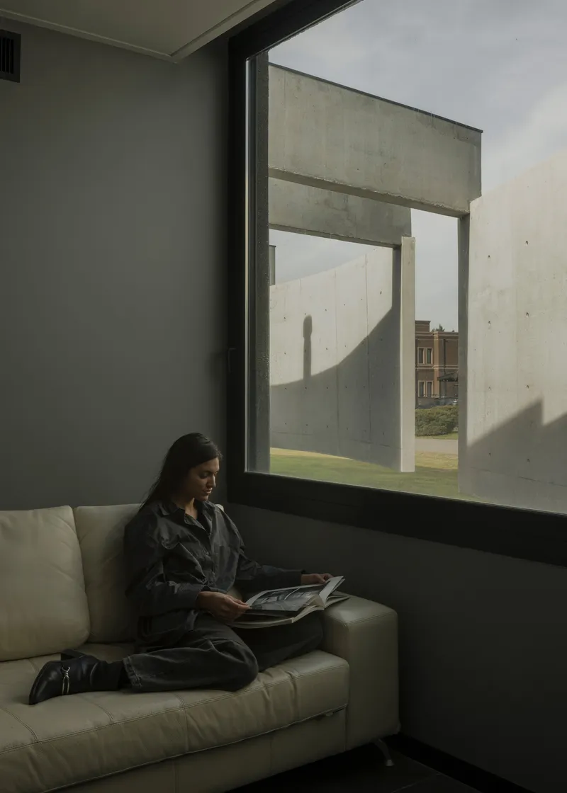 Interior reading nook with woman on sofa beside a deep-set window framing concrete courtyard walls