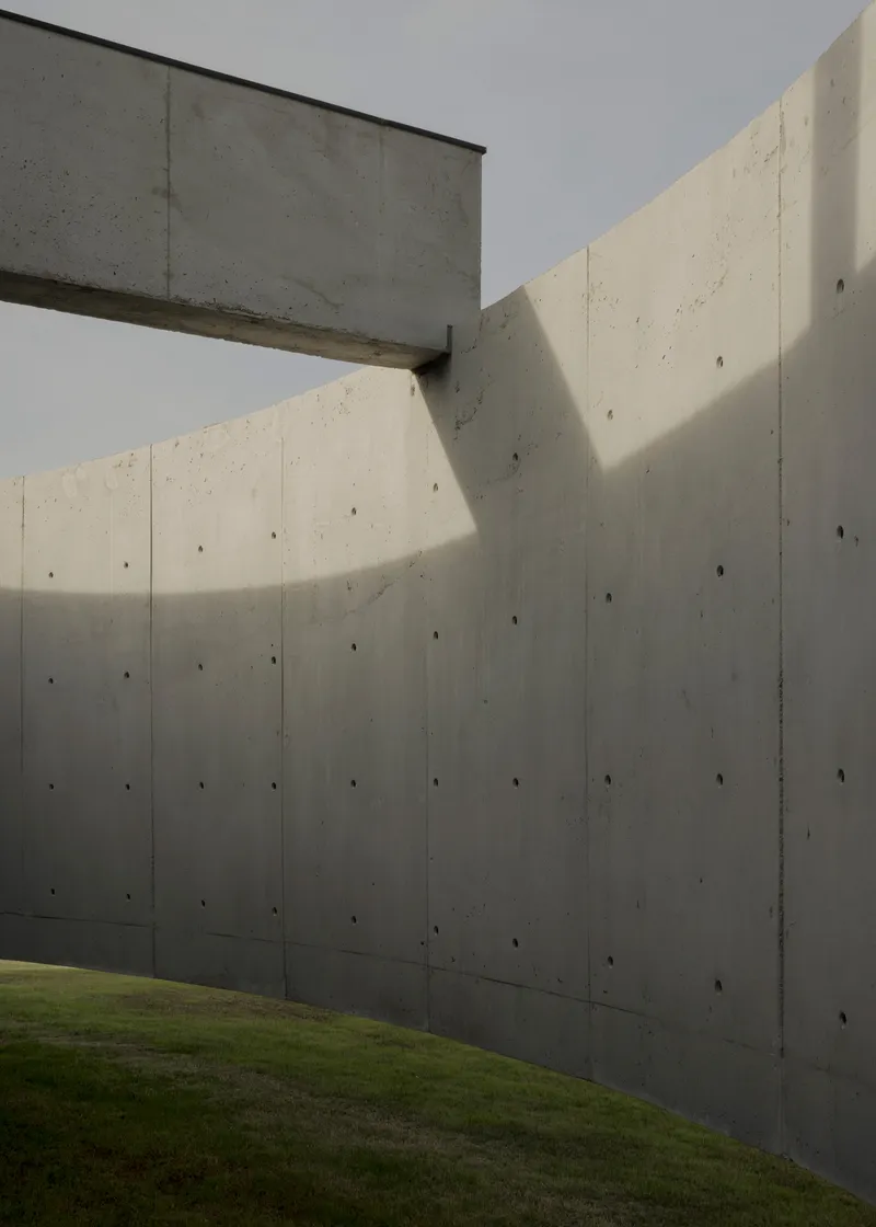 Boardformed concrete walls meeting at an angled corner with a cantilevered beam above green lawn