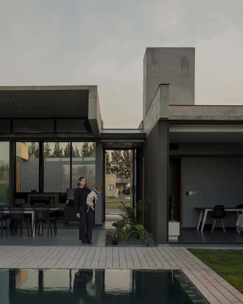 Courtyard with swimming pool and concrete tower viewed past a woman standing among potted plants
