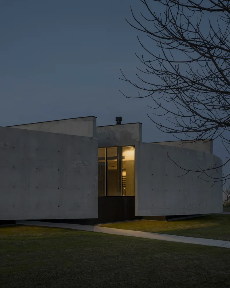 Dusk view of the illuminated corner window within the board-formed concrete facade beneath bare branches
