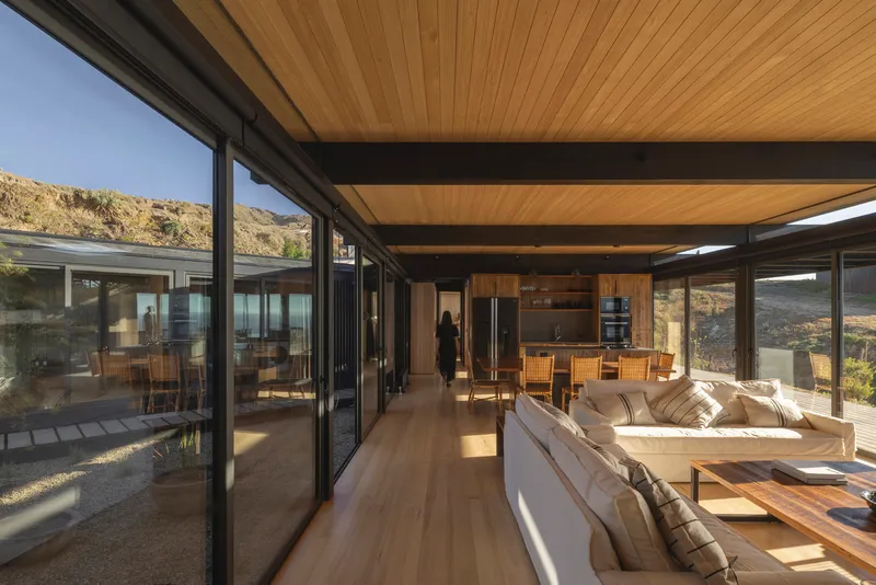 Open-plan living space with timber ceiling, exposed steel beams and floor-to-ceiling glass overlooking dry hillside